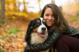Eine lächelnde junge Frau mit langen braunen Haaren, die eine schwarze Lederjacke trägt, hockt in einem herbstlichen Wald und umarmt liebevoll einen glücklichen Berner Sennenhund. Der Hund blickt mit offenem Mund direkt in die Kamera. Der Hintergrund zeigt ein unscharfes Meer aus gelben und orangefarbenen Herbstblättern. Dieses warme Porträt symbolisiert die starke Mensch-Hund-Bindung und das tiefe Vertrauen, das im Hundezentrum gelebt und gefördert wird.