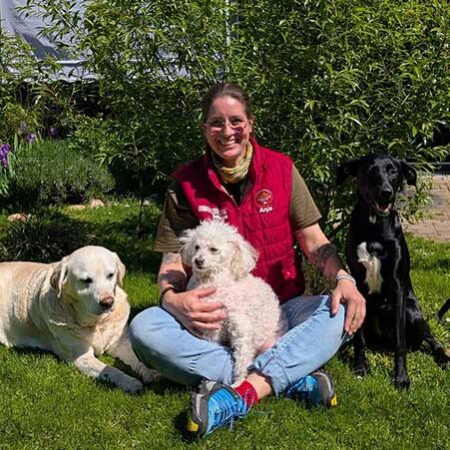 „Hundefachkraft Anja vom Hundezentrum Kuppenheim sitzt lachend im Garten auf einer sonnigen Wiese, umgeben von drei Hunden: einem ruhenden Labrador Retriever mit hellem Fell, einem kleinen weißen Pudel auf ihrem Schoß und einem großen, schwarzen Mischlingshund mit weißer Brust. Im Hintergrund blühen lila Iris und grüne Sträucher – eine harmonische Szene tiergestützter Arbeit im Freien.“