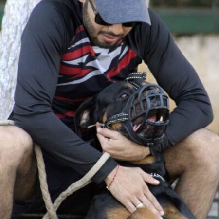 Ein bärtiger Mann in einer grauen Mütze, Sonnenbrille und einem langärmeligen Hemd im Motocross-Stil sitzt und hält einen Rottweiler liebevoll auf seinem Schoß. Der Hund liegt entspannt auf dem Rücken und trägt einen schwarzen Korbmaulkorb. Der Mann hat eine Hand sanft am Maulkorb und die andere am Bauch des Hundes. Die Interaktion zeigt Zuneigung und Vertrauen trotz des Maulkorbs. Der Hintergrund ist ein leicht unscharfer Park- oder Außenbereich mit Baumstämmen und Vegetation. Eine dicke, geflochtene Seilleine liegt neben ihnen. Nahaufnahme, Tageslicht.